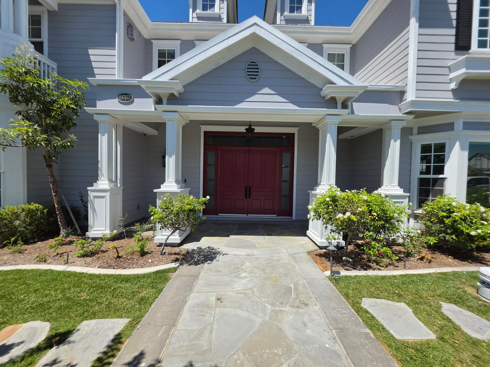 Gray house with white columned portico, burgundy double doors, flagstone walkway, and landscape plantings