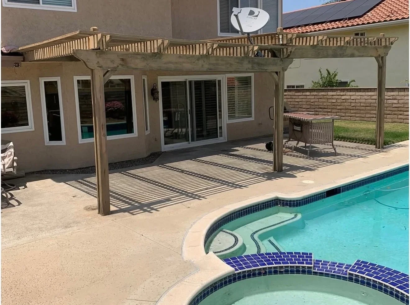 Wood pergola with slatted roof over concrete patio beside swimming pool with spa